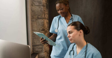 Duas profissionais de enfermagem com estetoscópio analisam informações no computador