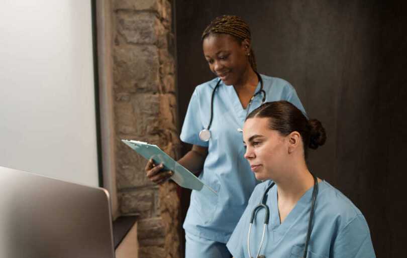 Duas profissionais de enfermagem com estetoscópio analisam informações no computador
